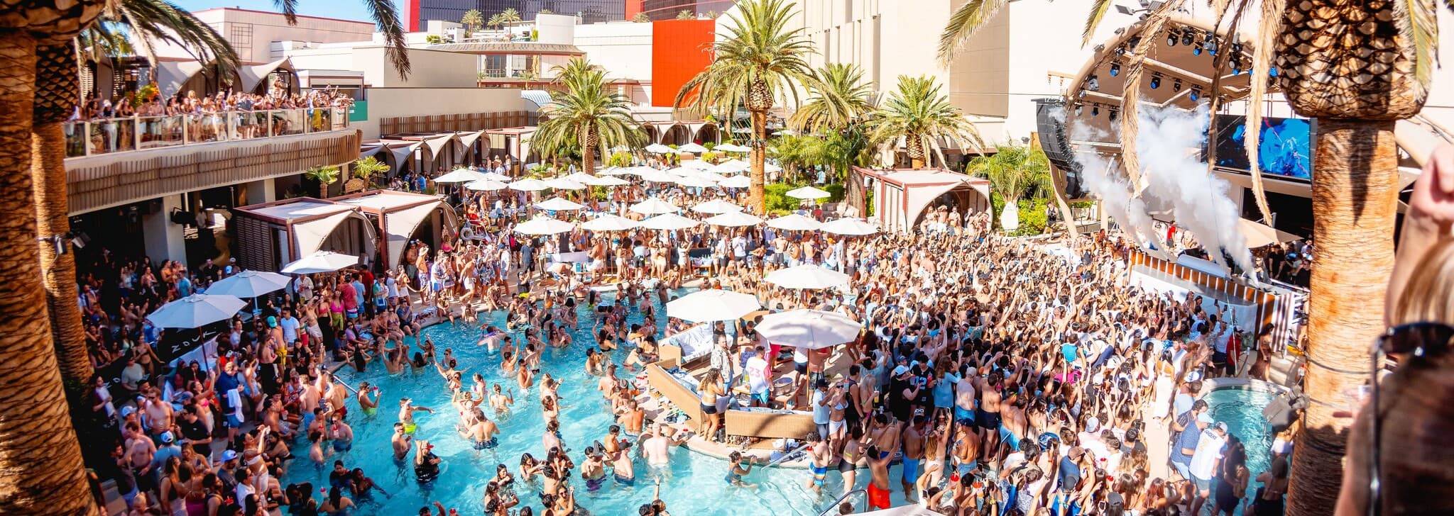 Large crowd gathered at a Las Vegas dayclub pool party featuring palm trees, poolside cabanas, DJs performing on stage, and guests dancing in and around the swimming pool.
